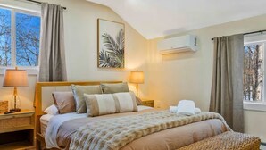 Ein Schlafzimmer mit einem Bett, Nachttischlampen, einer Bank, einer Wand-Klimaanlage und einem Fenster mit Vorhängen.