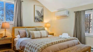 A bedroom with a bed, bedside lamps, a bench, a wall-mounted air conditioner, and a window with curtains.
