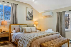 Ein Schlafzimmer mit einem Bett, Nachttischlampen, einer Bank, einer Wand-Klimaanlage und einem Fenster mit Vorhängen.