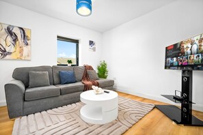 Ein modernes Wohnzimmer mit einem grauen Sofa, einem weißen Couchtisch, einem Flachbildfernseher auf einem Standfuß und einem Fenster mit Jalousien.