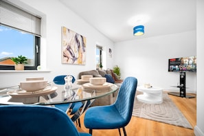 Una sala moderna con una mesa de comedor de cristal, sillas azules, un sofá, un televisor y obras de arte en la pared.