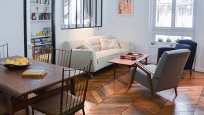 Un salon comprenant un canapé, une table à manger avec des chaises, une bibliothèque et une fenêtre donnant sur des immeubles.