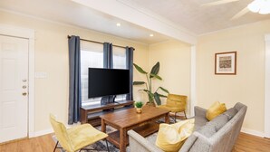 Een woonkamer met een televisie, een houten salontafel, gele fauteuils, een grijze bank en een plant.