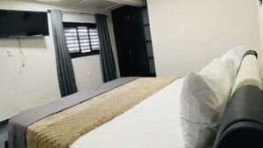 A room with a bed, a flat-screen TV mounted on the wall, a window with blinds, and a door.