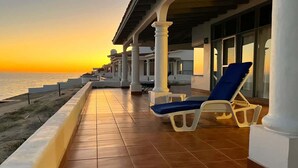 Una terraza con columnas blancas, sillones azules y vista al mar al atardecer.