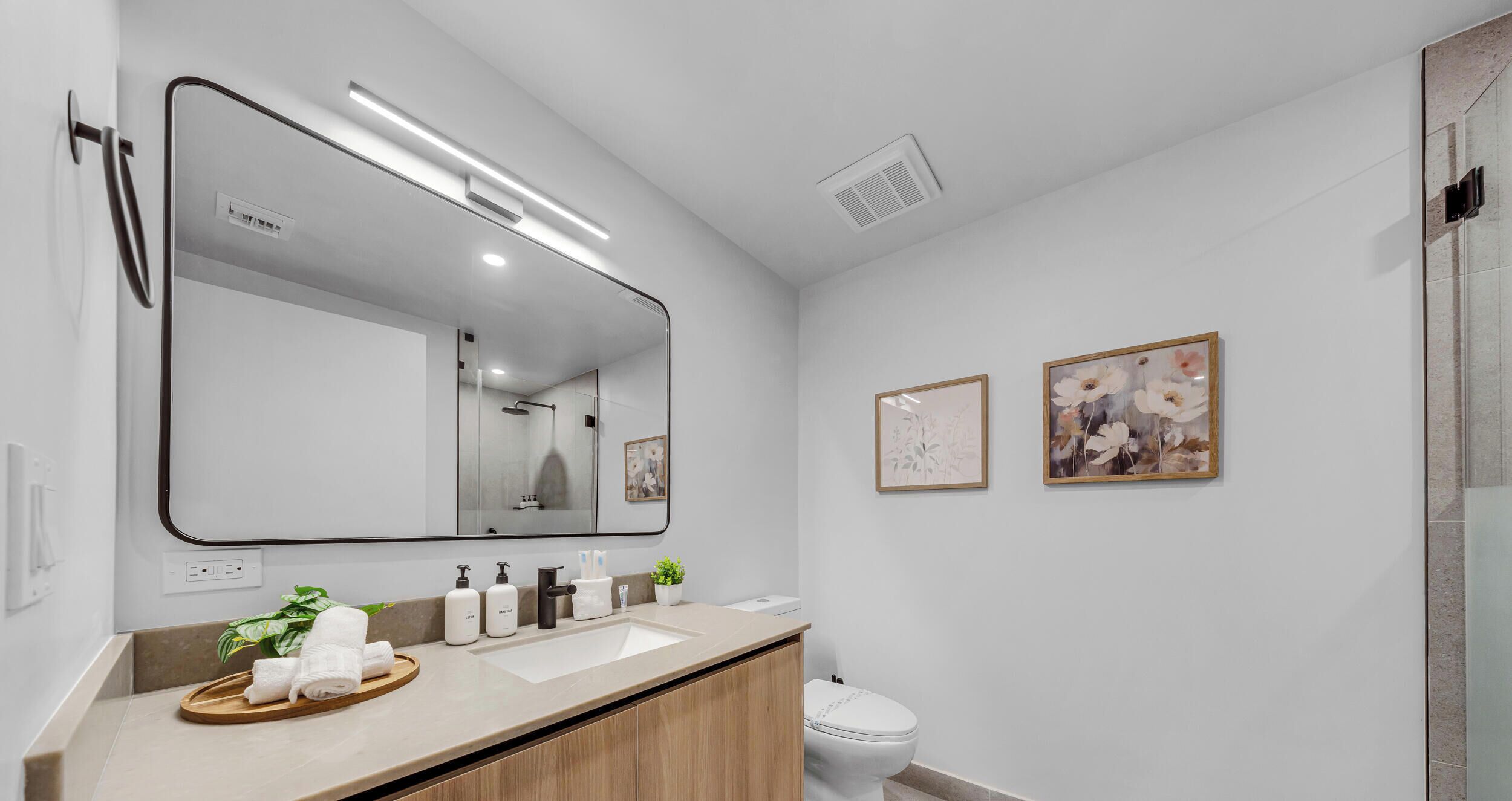 A modern bathroom with a large mirror, a sink, a toilet, and framed artwork on the wall.