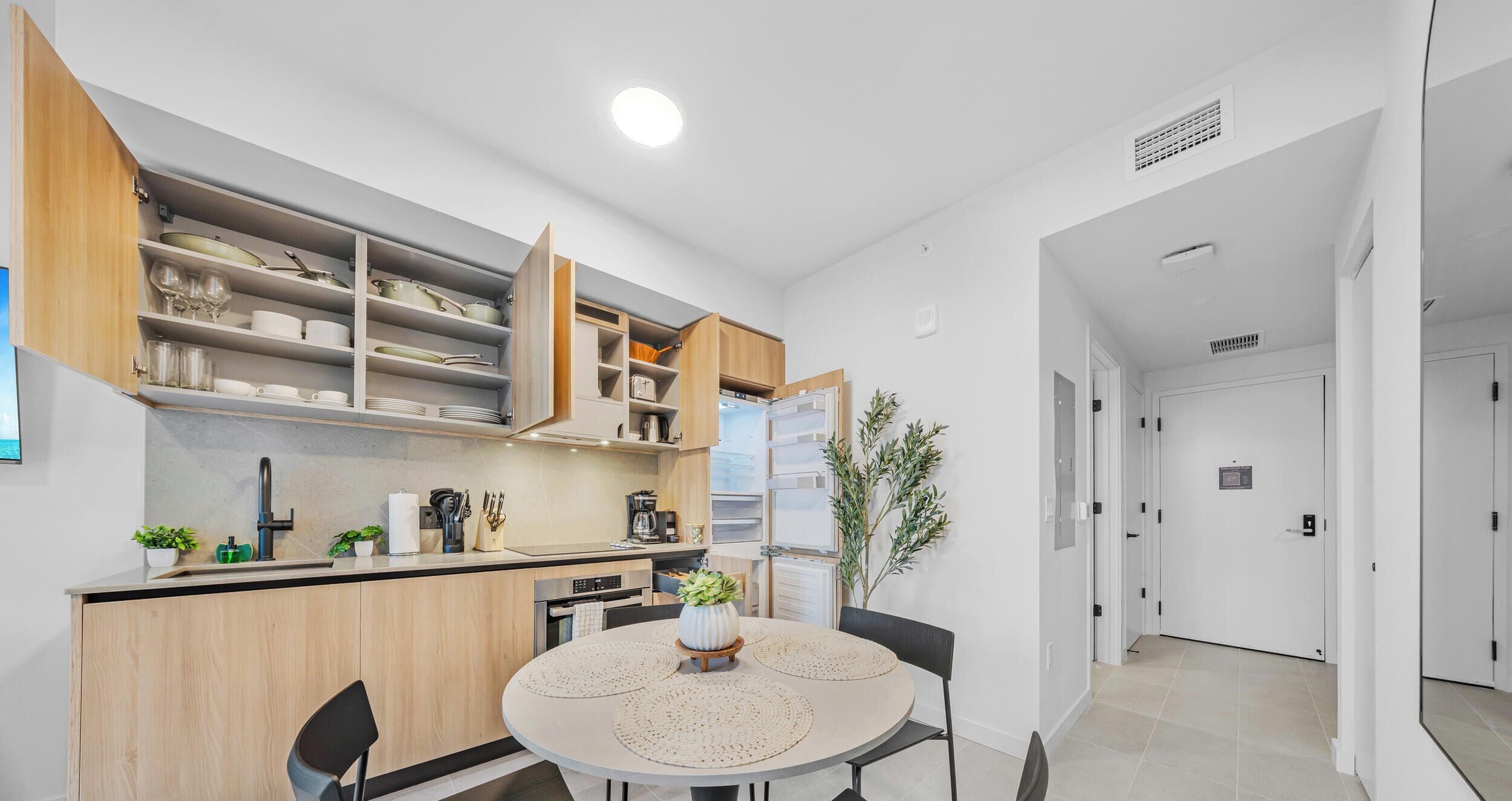 Cocina compacta con armarios abiertos, una mesa de comedor con individuales y una planta en maceta.