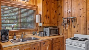 A wooden cabin kitchen with a stove, microwave, toaster, sink, and hanging utensils. - Lazy Trout Spruce Cabin (Greer)