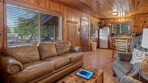 A wooden interior with a leather sofa, a wicker chair, a dining table, and a kitchen area. - Lazy Trout Spruce Cabin (Greer)