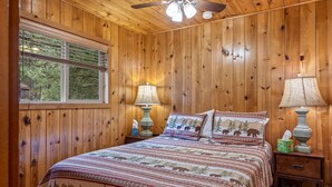 A wooden-paneled room with a bed, two bedside lamps, a ceiling fan, and a window with blinds. - Lazy Trout Spruce Cabin (Greer)