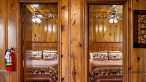 A mirrored room with wooden walls, a ceiling fan, a fire extinguisher, and a bed with a forest-themed bedspread. - Lazy Trout Aspen Cabin (Greer)
