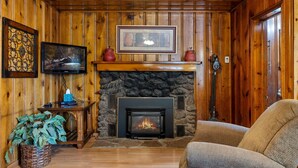 A cozy room with a stone fireplace, wooden walls, a television, and a comfortable chair. - Lazy Trout Aspen Cabin (Greer)