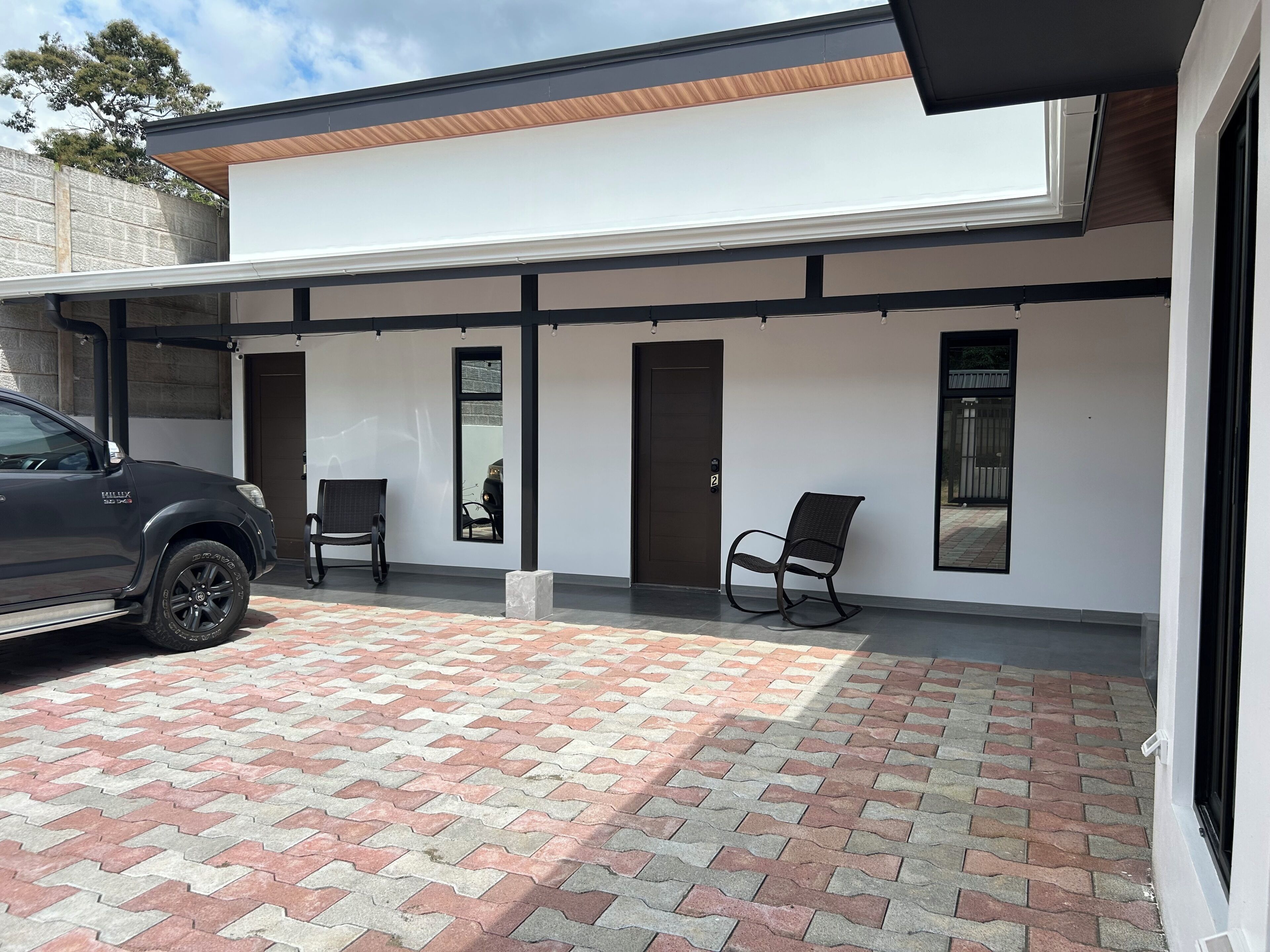 Un edificio moderno con un patio techado, dos sillas y un vehículo estacionado.