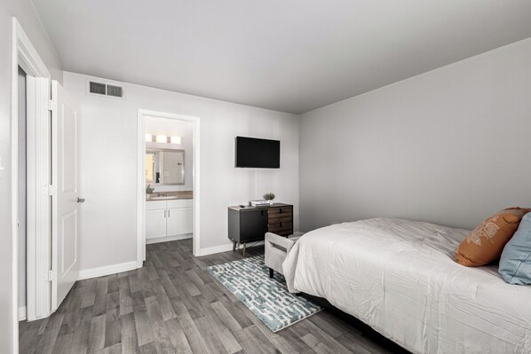 Un dormitorio con cama, mesita de noche, televisor, cómoda y baño con espejo y lavamanos.