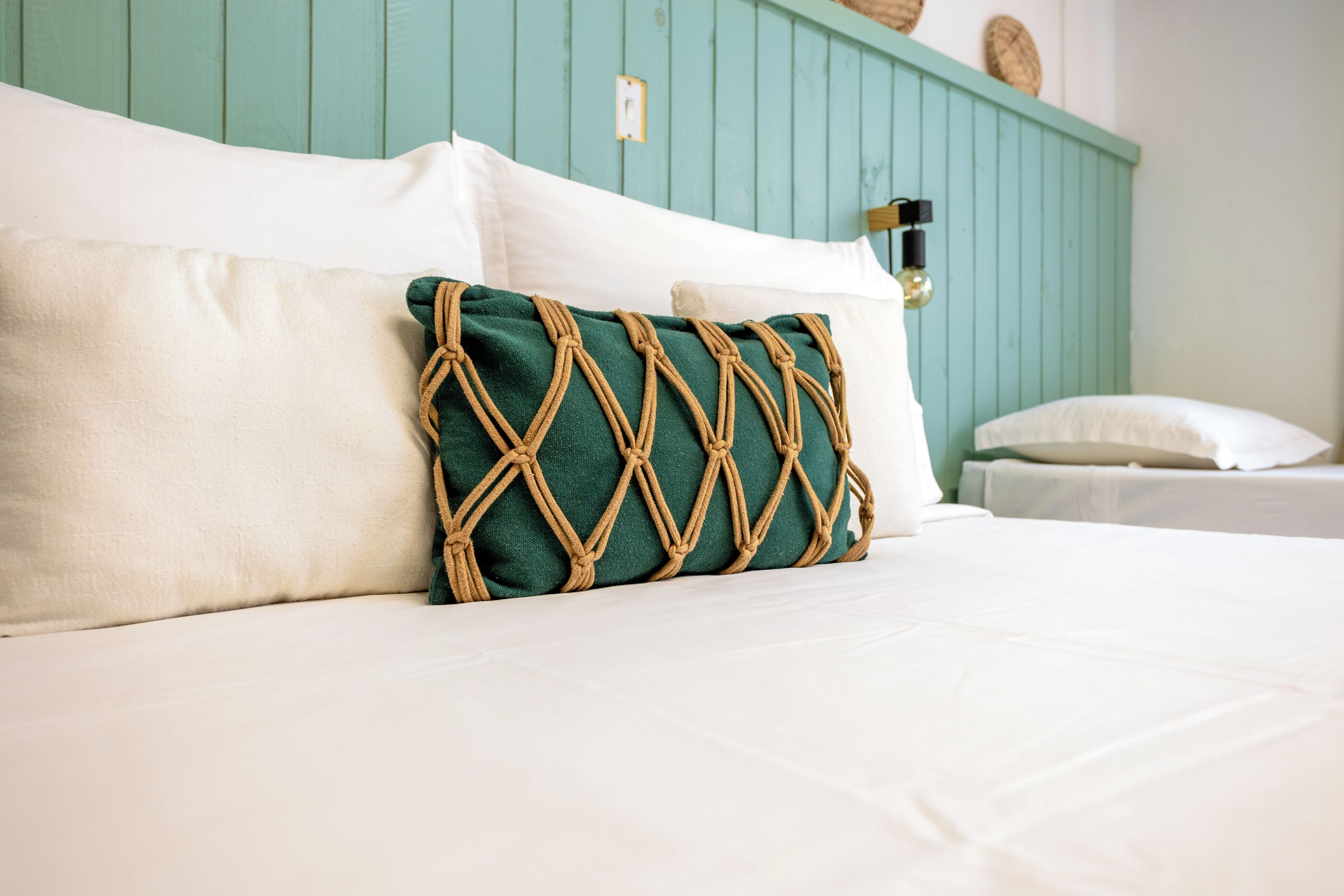 A bed with white linens, a green macrame pillow, and a teal headboard.
