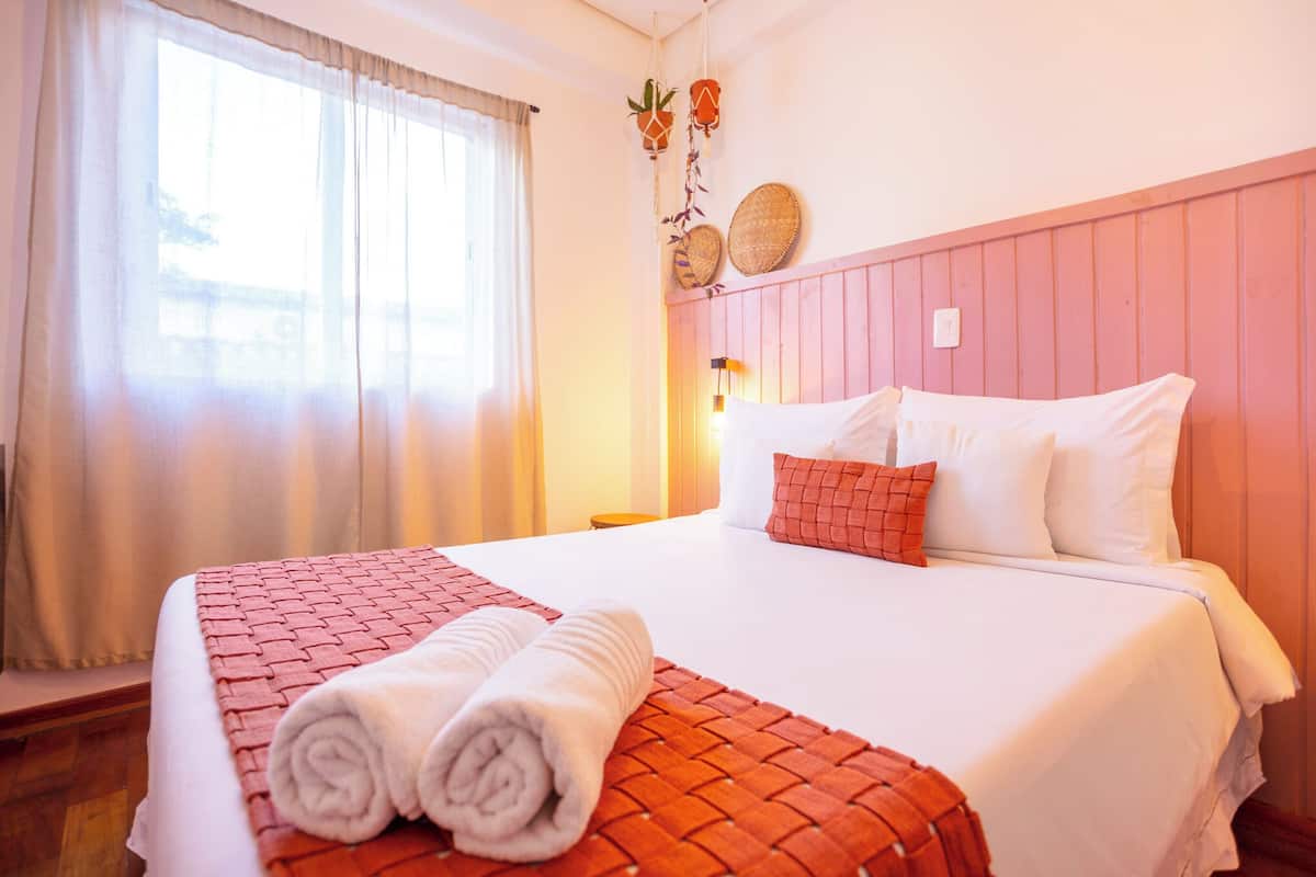 A hotel room with a bed, a window with curtains, a wall-mounted lamp, and decorative items.