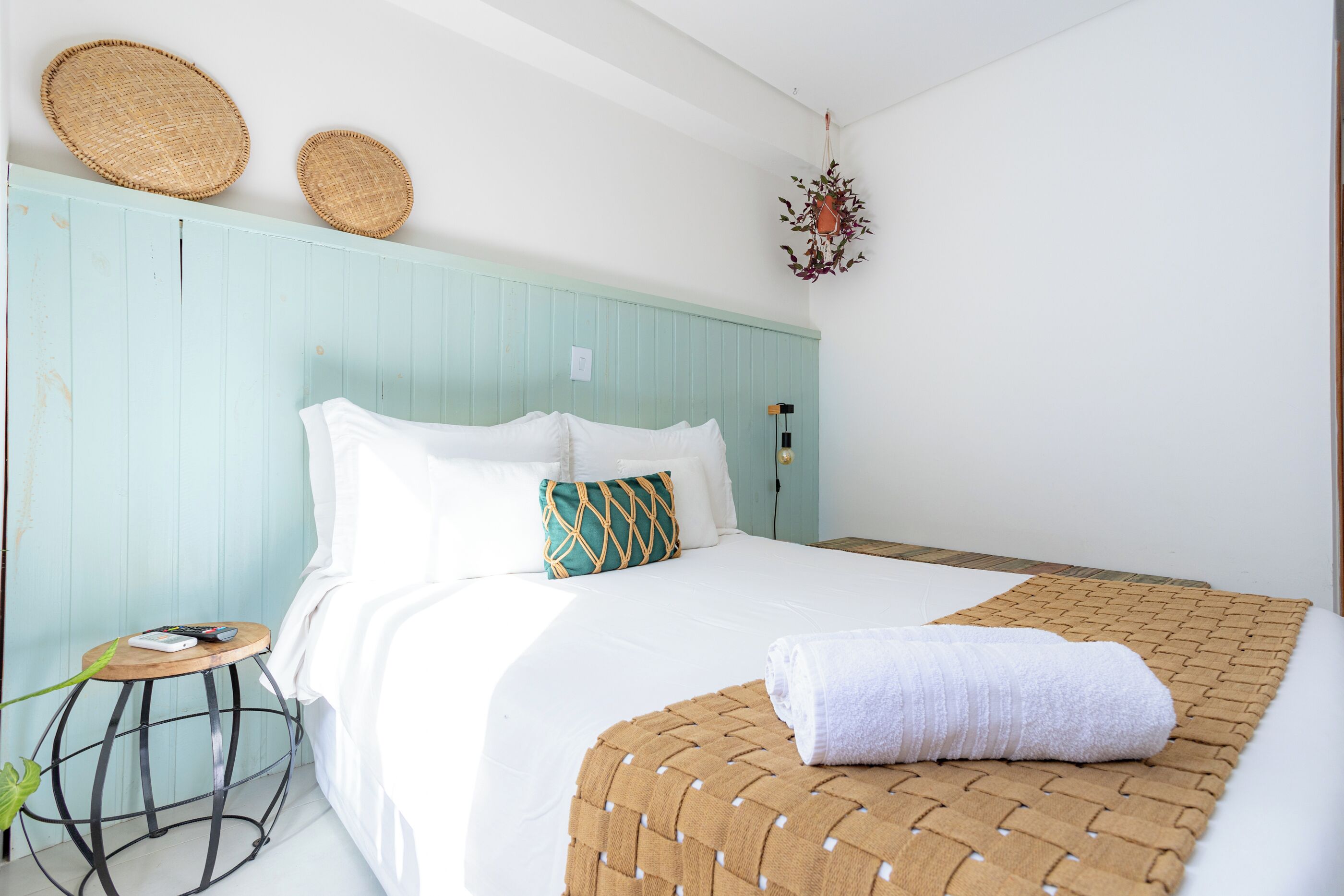 Een bed met witte beddengoed, een geweven plaid en een klein rond tafeltje met een plant.