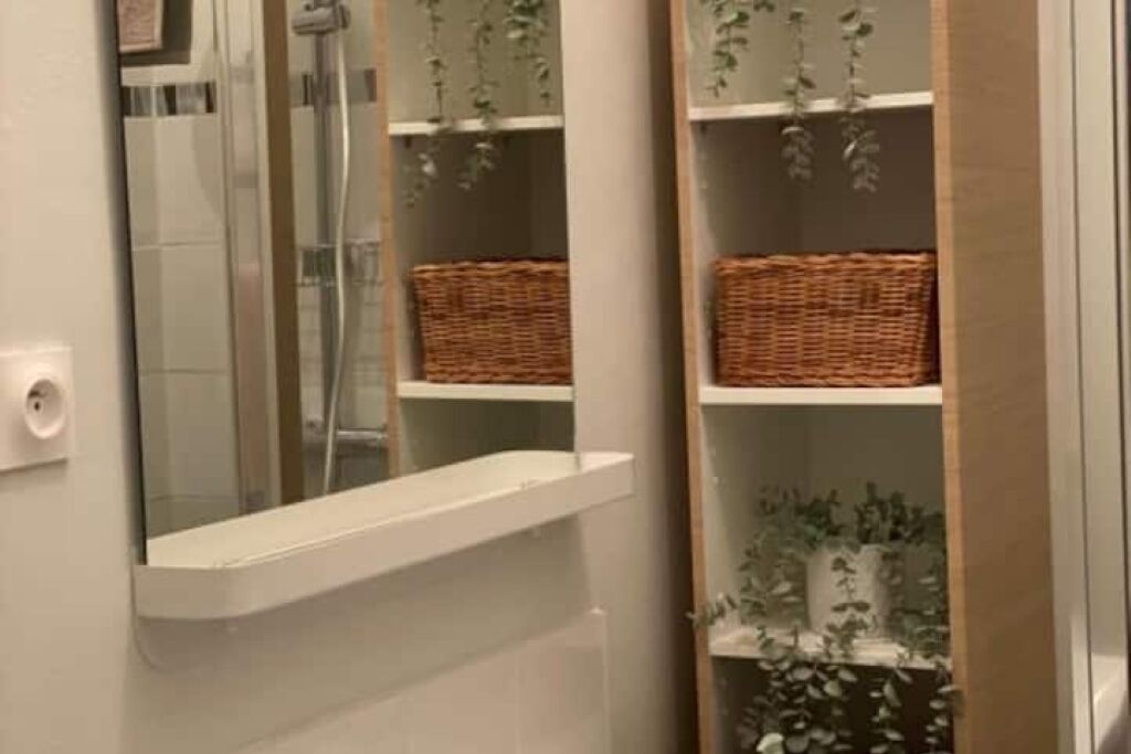Une salle de bain dotée d'une douche à parois de verre, d'étagères blanches ornées de paniers en osier et d'un miroir qui agrandit l'espace.