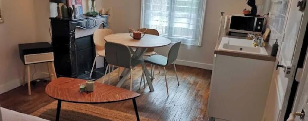 Een compacte keuken met een eettafel en stoelen, een kleine koelkast en een raam met zonwering.