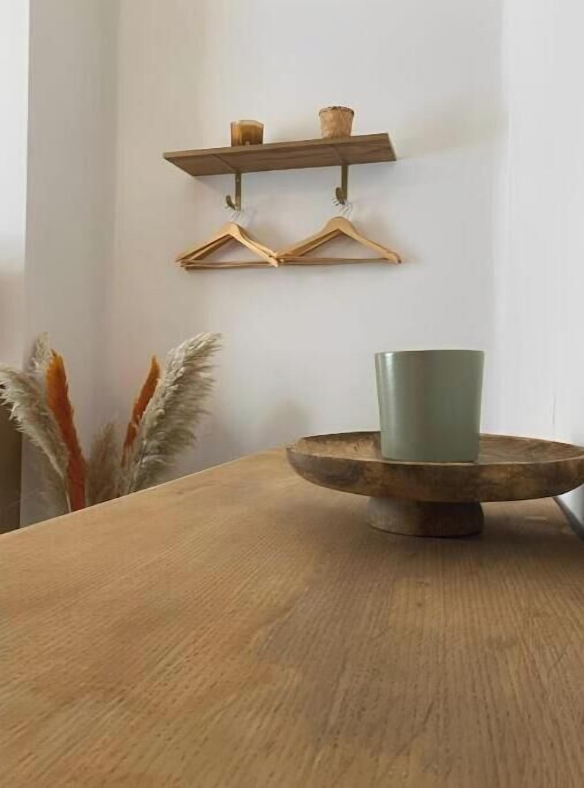 A wooden table with a green cup, a wooden bowl, and a wall-mounted shelf with two wooden hangers and two candles.