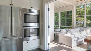 A modern kitchen with stainless steel appliances, a white dining area with a sofa, and large windows. - Luxury on Glen Lake (Eagle Lake)