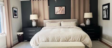 A bedroom with a large bed, two bedside tables with lamps, a nightstand, a window with curtains, and a painting on the wall.