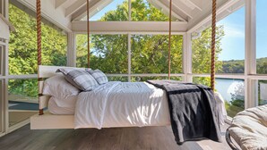 A bedroom with a hanging bed, large windows, and a view of trees and water. - Luxury on Glen Lake (Eagle Lake)