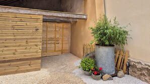 Una puerta de madera, una planta grande en maceta, una maceta pequeña con flores rojas y un barril de metal.