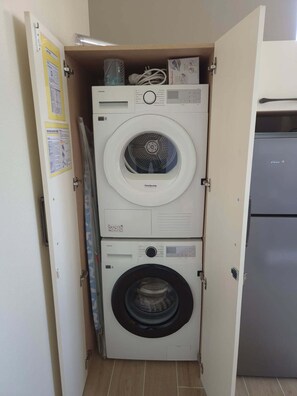 Une petite buanderie équipée de deux machines à laver et deux sèche-linge superposés, d’un réfrigérateur et d’une porte ornée d’affiches d’information.