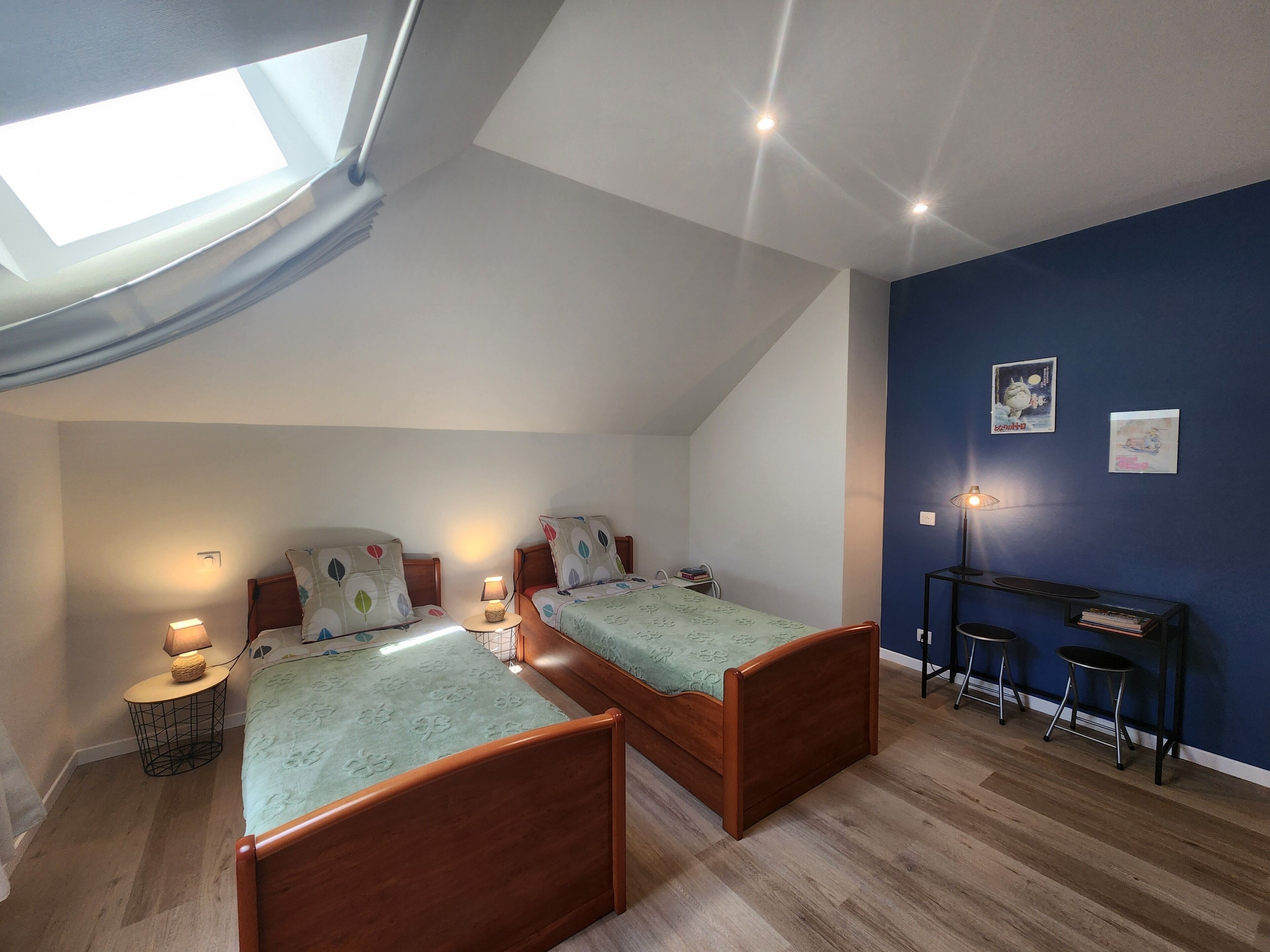 Une chambre avec deux lits en bois, une lucarne, un bureau avec une lampe et un mur d’accent bleu.