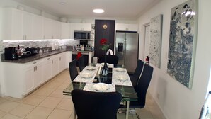 A modern kitchen with white cabinets, a microwave, and a refrigerator. There is a dining area with a glass table and chairs. - Cozy vacation home w/ a spacious outdoor area. Near to great beaches and casinos (Hollywood)