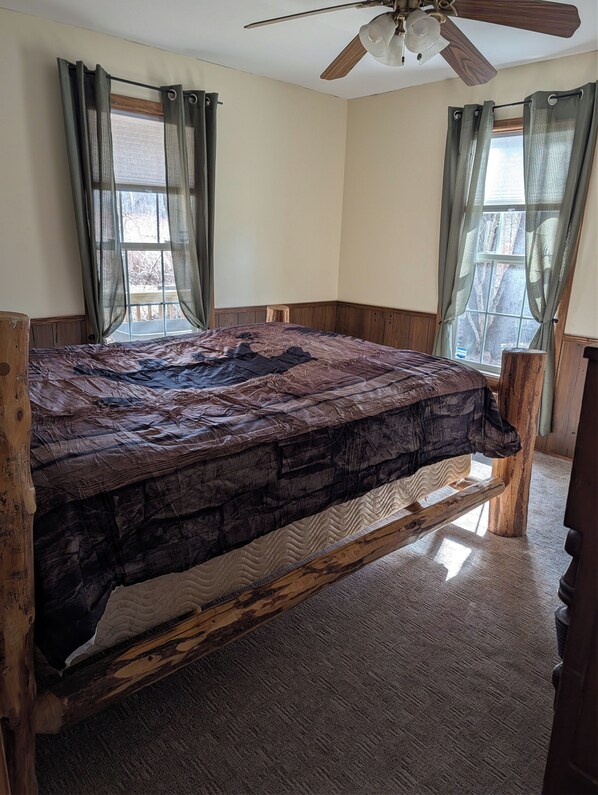 A bed with a wooden frame and a textured mattress, a ceiling fan, and two windows with curtains. - Fall is around the corner. Adventure is waiting for guests to explore. (Orangeville)
