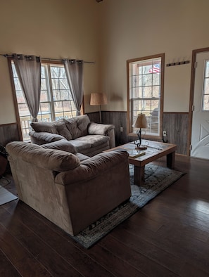 A living room with a brown sofa, a wooden coffee table, and a lamp. There are windows with curtains, a door, and a wall-mounted lamp.