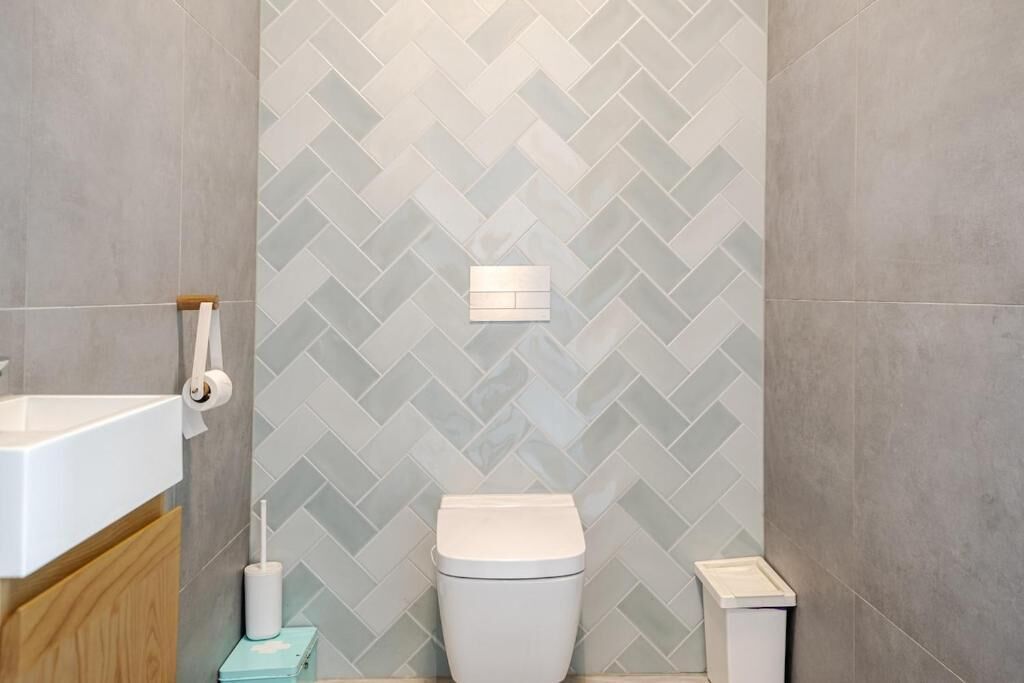 Un baño con salpicadero de azulejos tipo espiga, un inodoro, un lavabo y un cubo de basura.