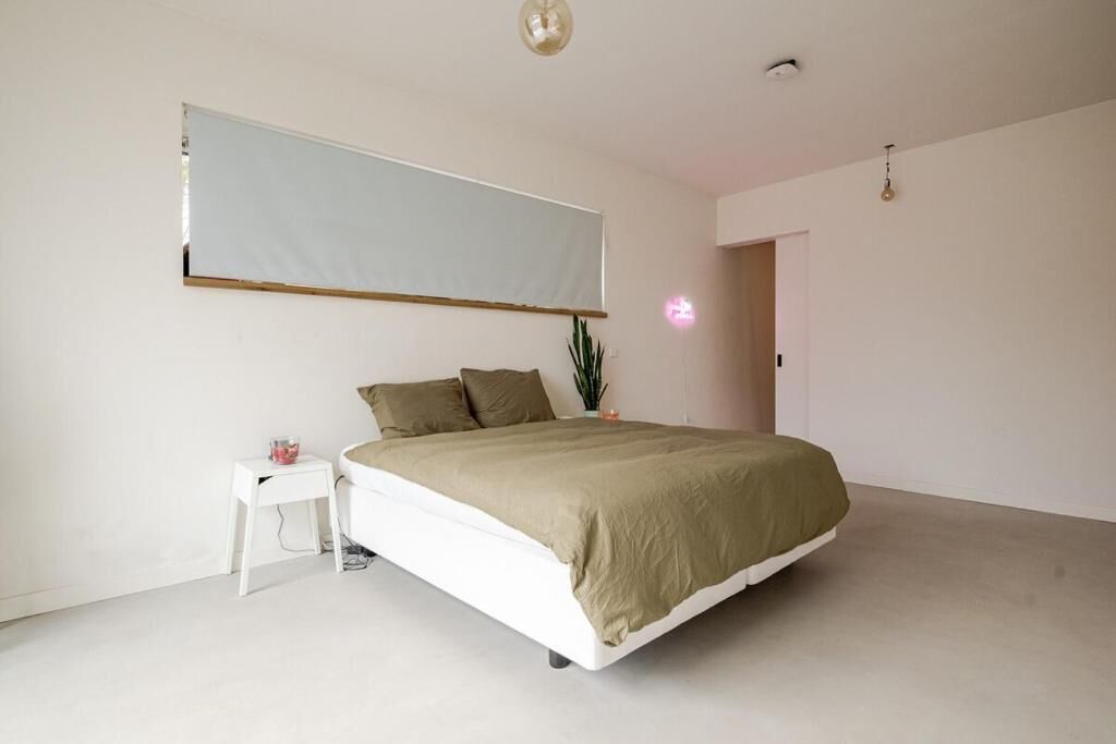 A bedroom with a bed, a small white table, a potted plant, and a large window.