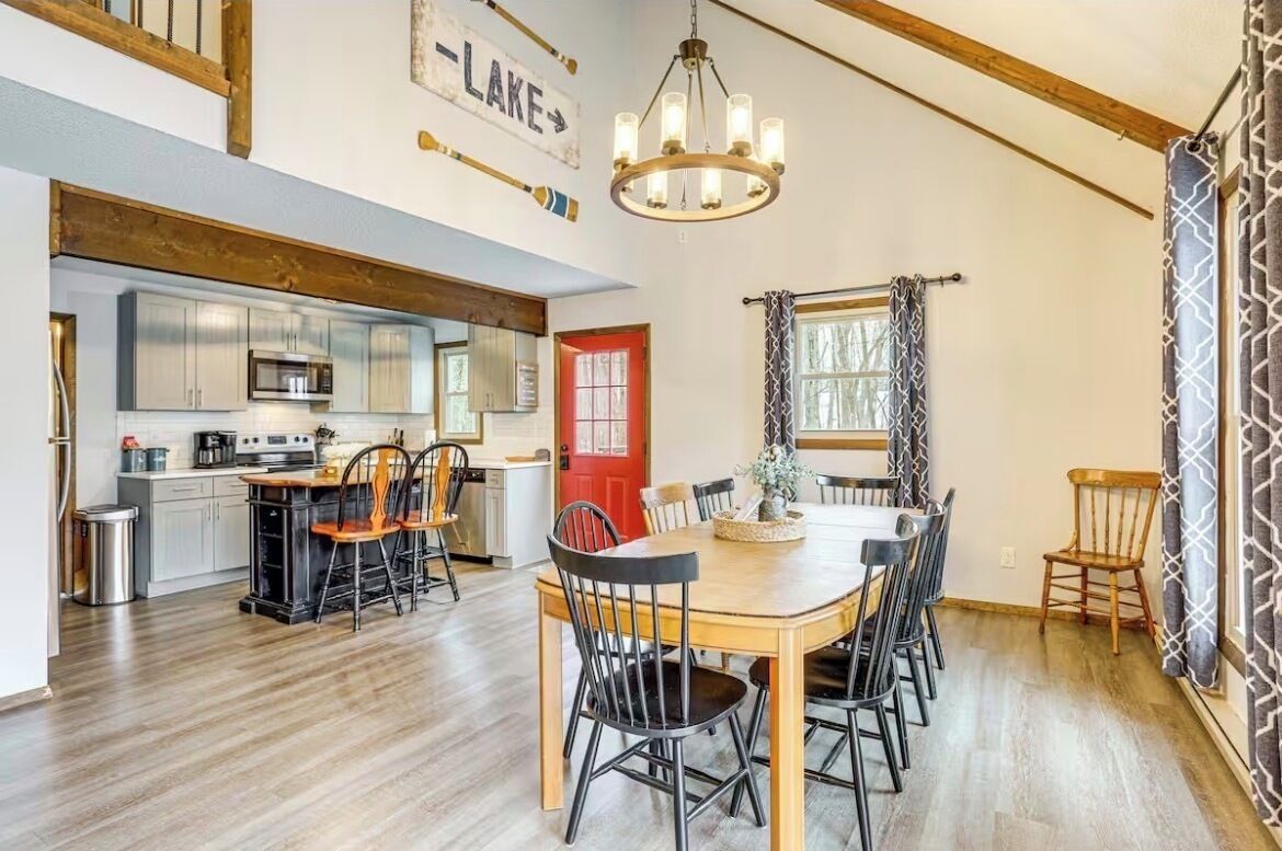 A dining area with a wooden table and chairs, a kitchen with modern appliances, and a red door.