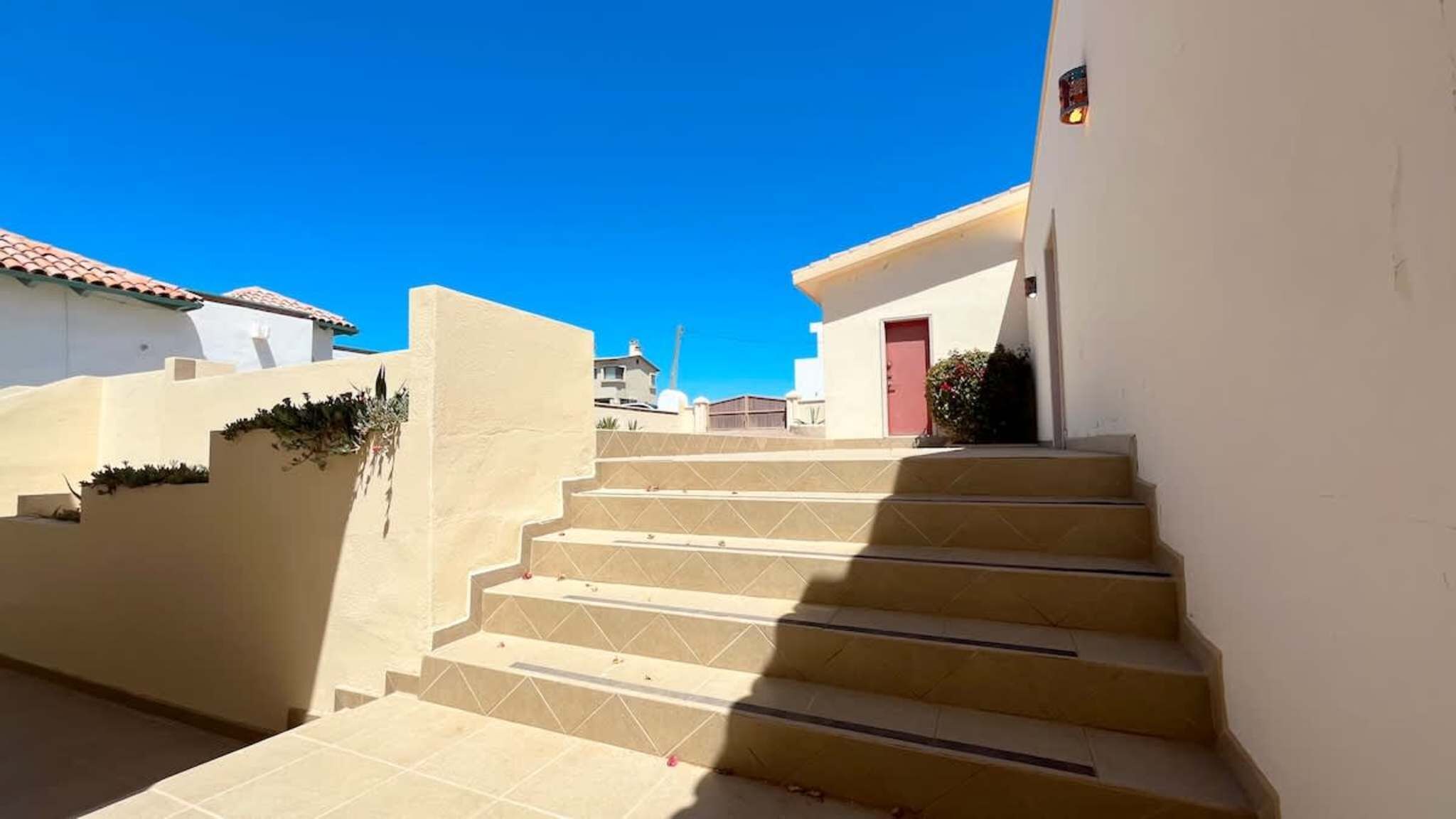 A set of beige stairs leading to a building with a red door, surrounded by white walls and a clear blue sky.