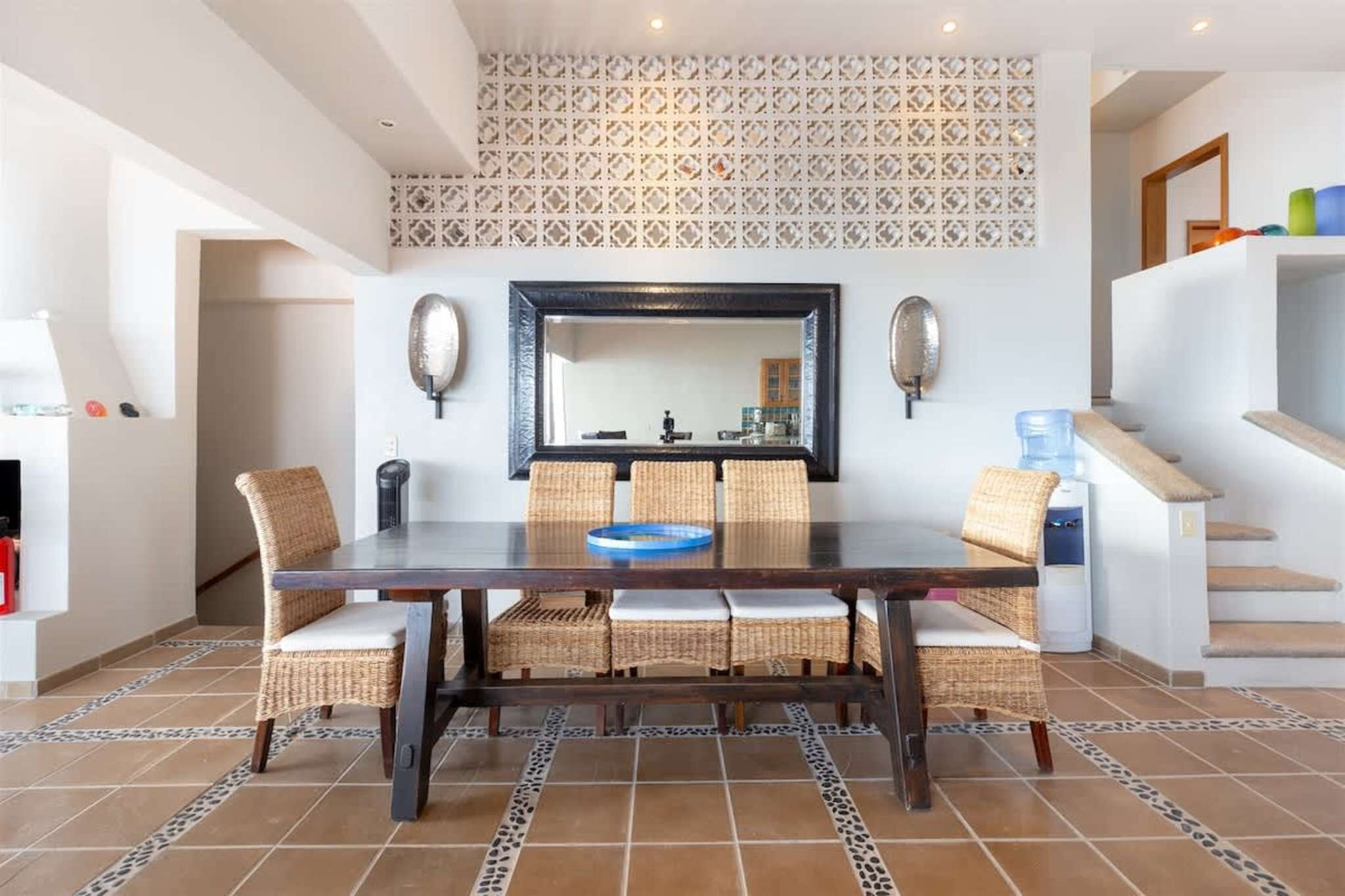 A dining area with a rectangular table, six wicker chairs, a large mirror, and a patterned wall above.