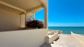 Ein Balkon mit Blick aufs Meer, eine Blumenkiste mit roten Blüten und ein strahlend blauer Himmel.