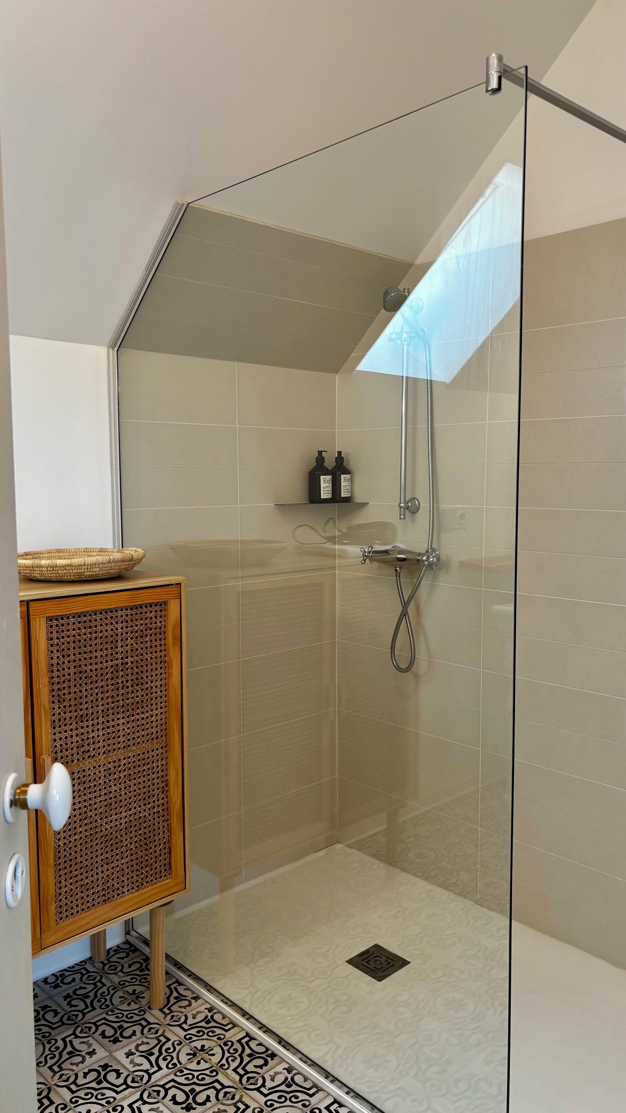 A glass-enclosed shower with a handheld showerhead, a wooden cabinet with a woven door, and a patterned floor.