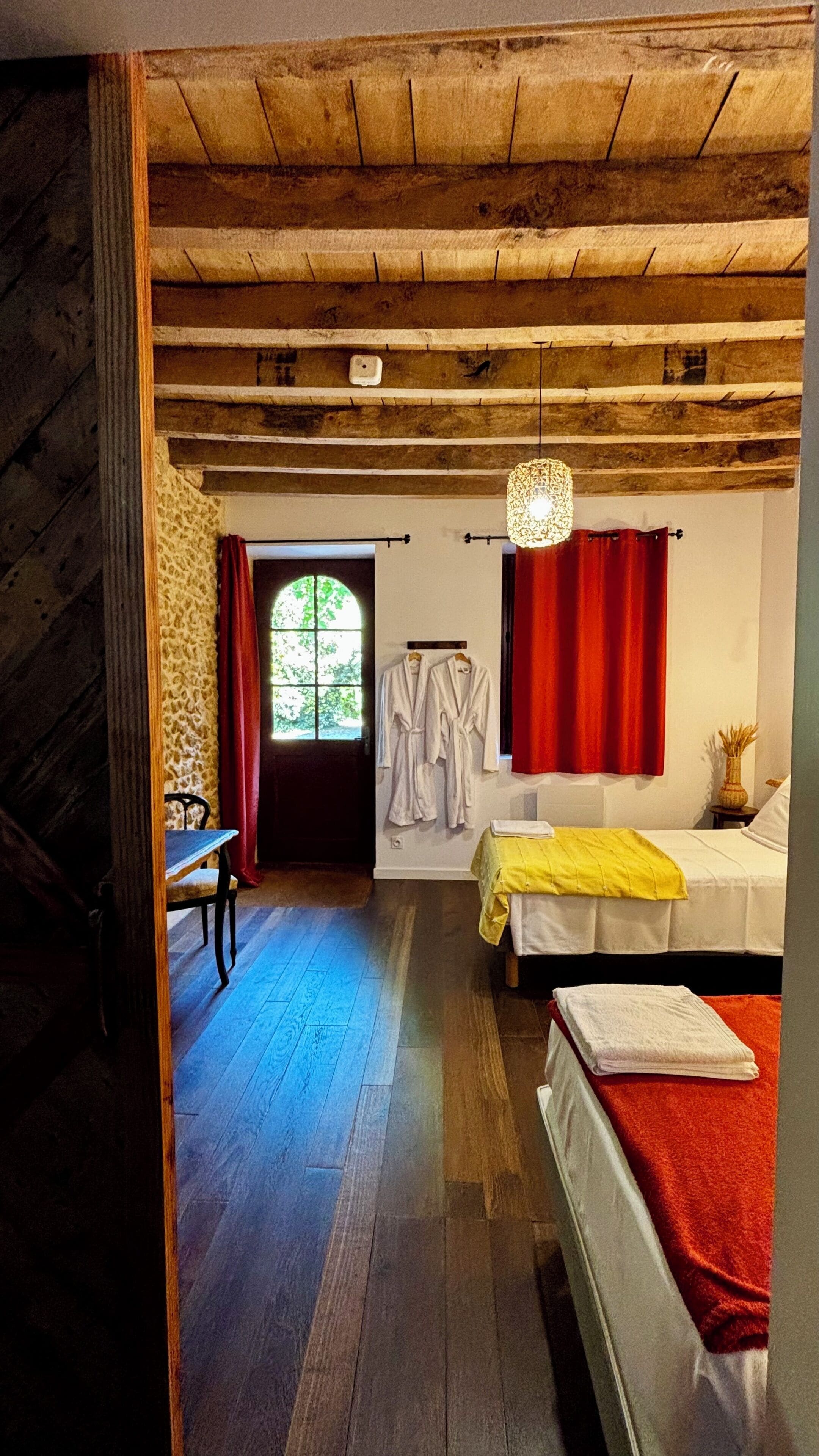 Una habitación con techo de madera, una cama con manta roja, una manta amarilla en otra cama, una ventana con cortinas rojas y una silla.