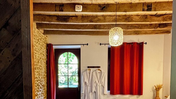 A room with a wooden ceiling, a bed with a red blanket, a yellow blanket on another bed, a window with red curtains, and a chair. - LE DOMAINE DU PETIT TERTRE (Sainte-Sabine-sur-Longève)