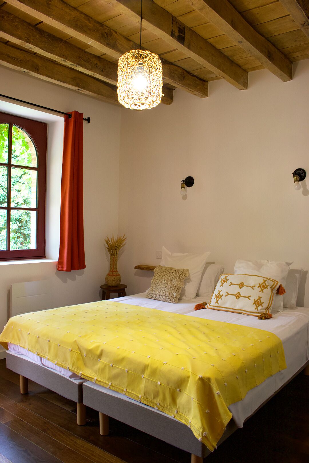 Um quarto com teto de madeira, uma cama com colcha amarela, uma janela com cortinas vermelhas e um lustre.