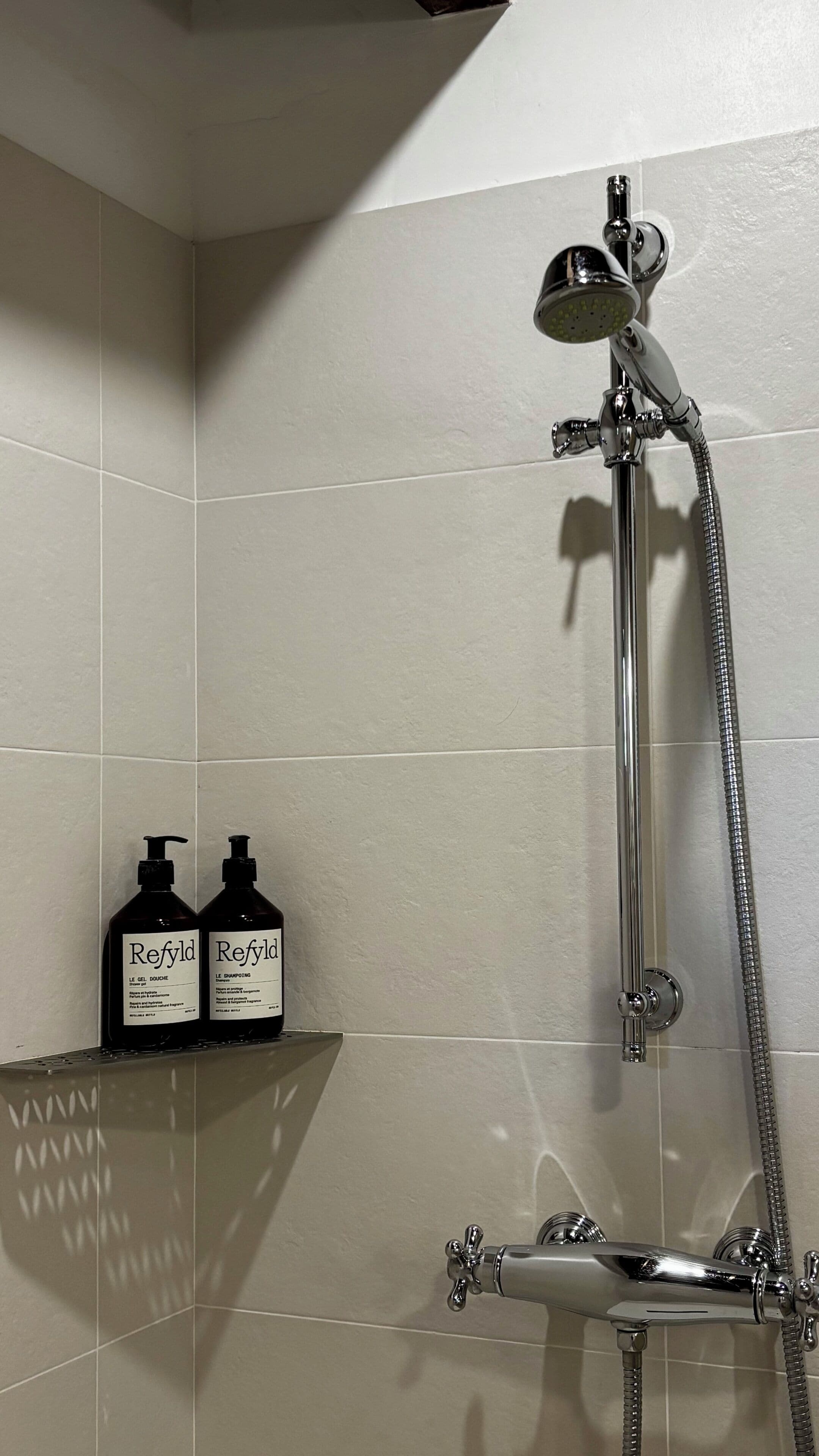 Baño con ducha, dos botellas de champú y gel Refyld, y pared de azulejos.
