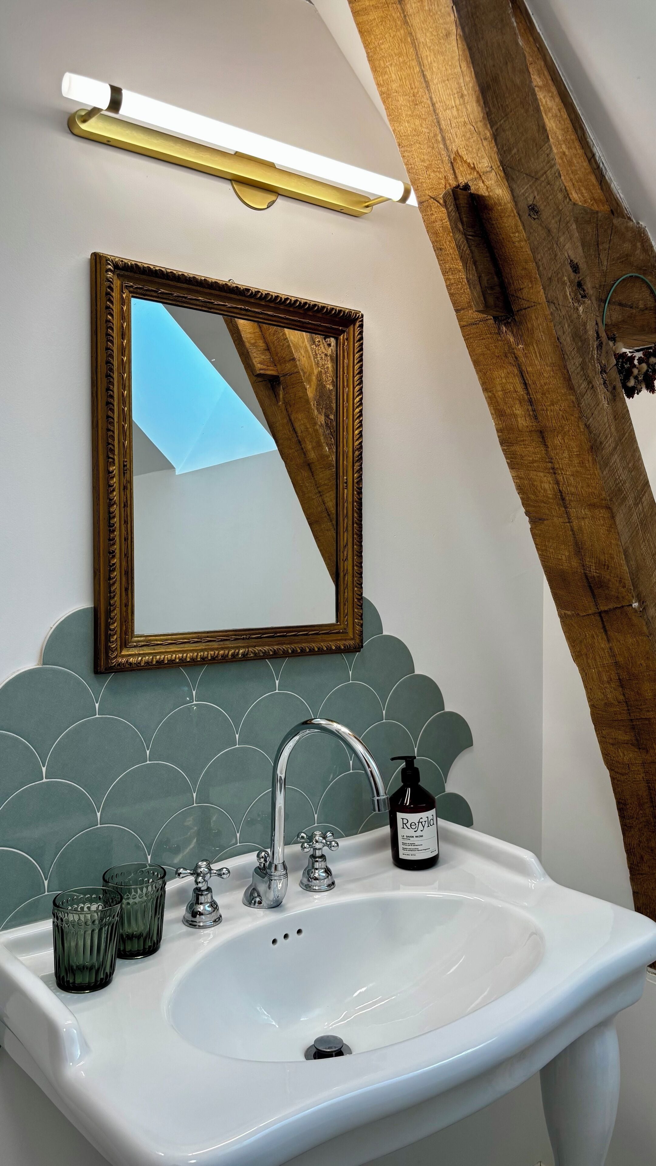 Un baño con un lavamanos blanco, un espejo con marco dorado, una pared con azulejos verdes y una viga de madera.