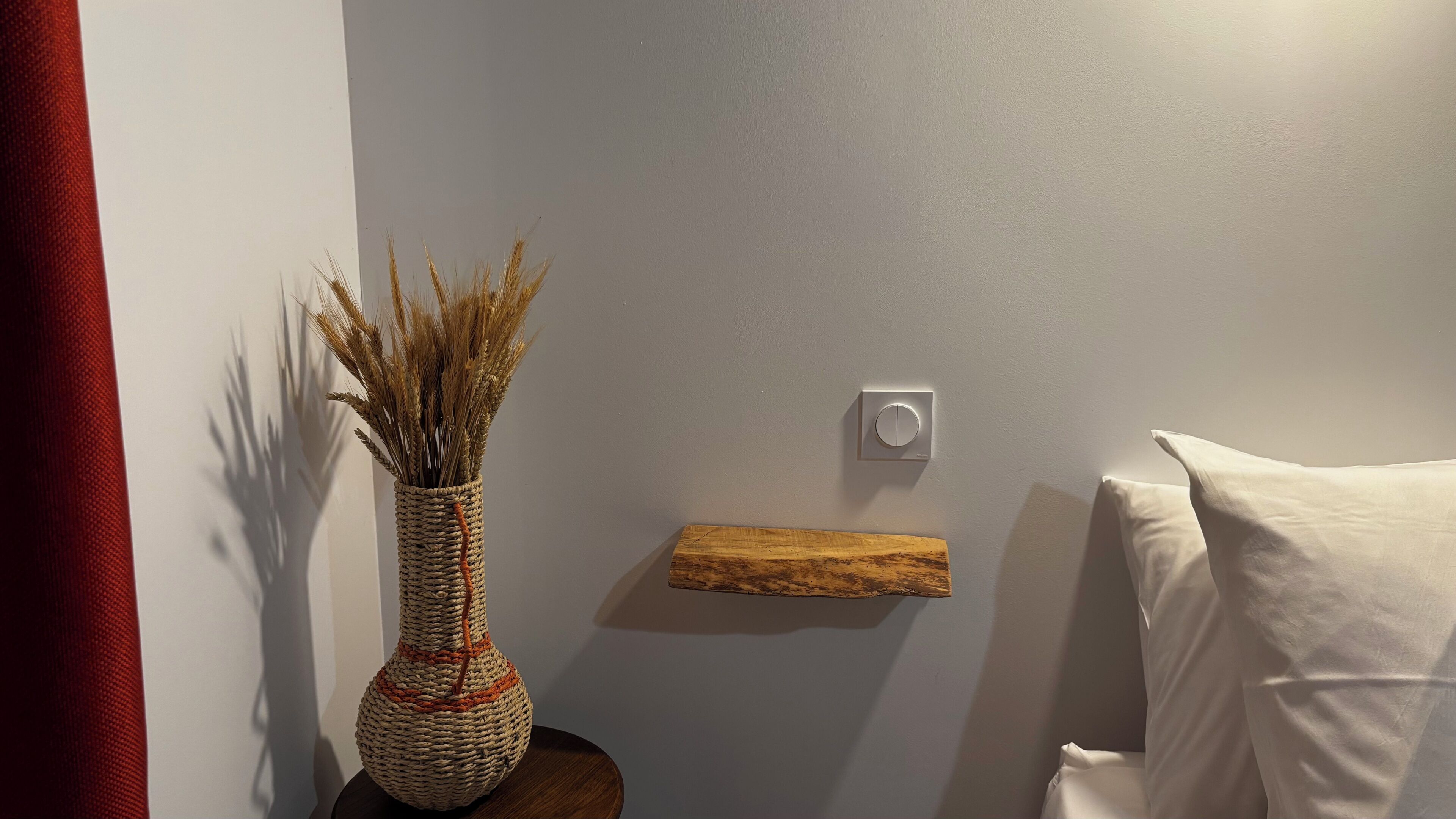 A room with a woven vase with dried plants on a wooden side table, a thermostat on the wall, and a white pillow on a bed.