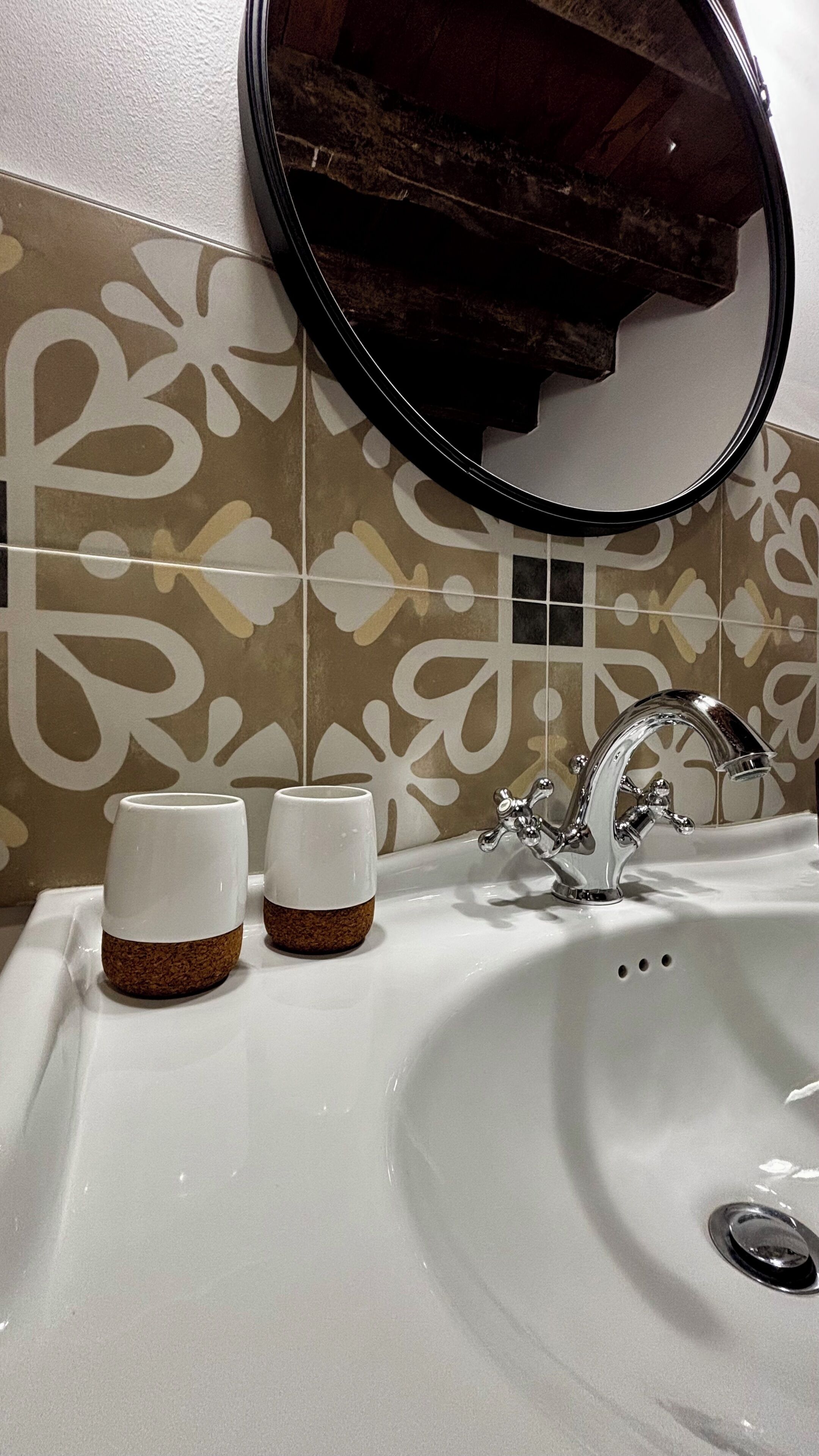 Een badkamer met een wit wastafel, twee witte bekers met kurken onderzetters, een zilveren kraan en een spiegel met een bruine lijst.