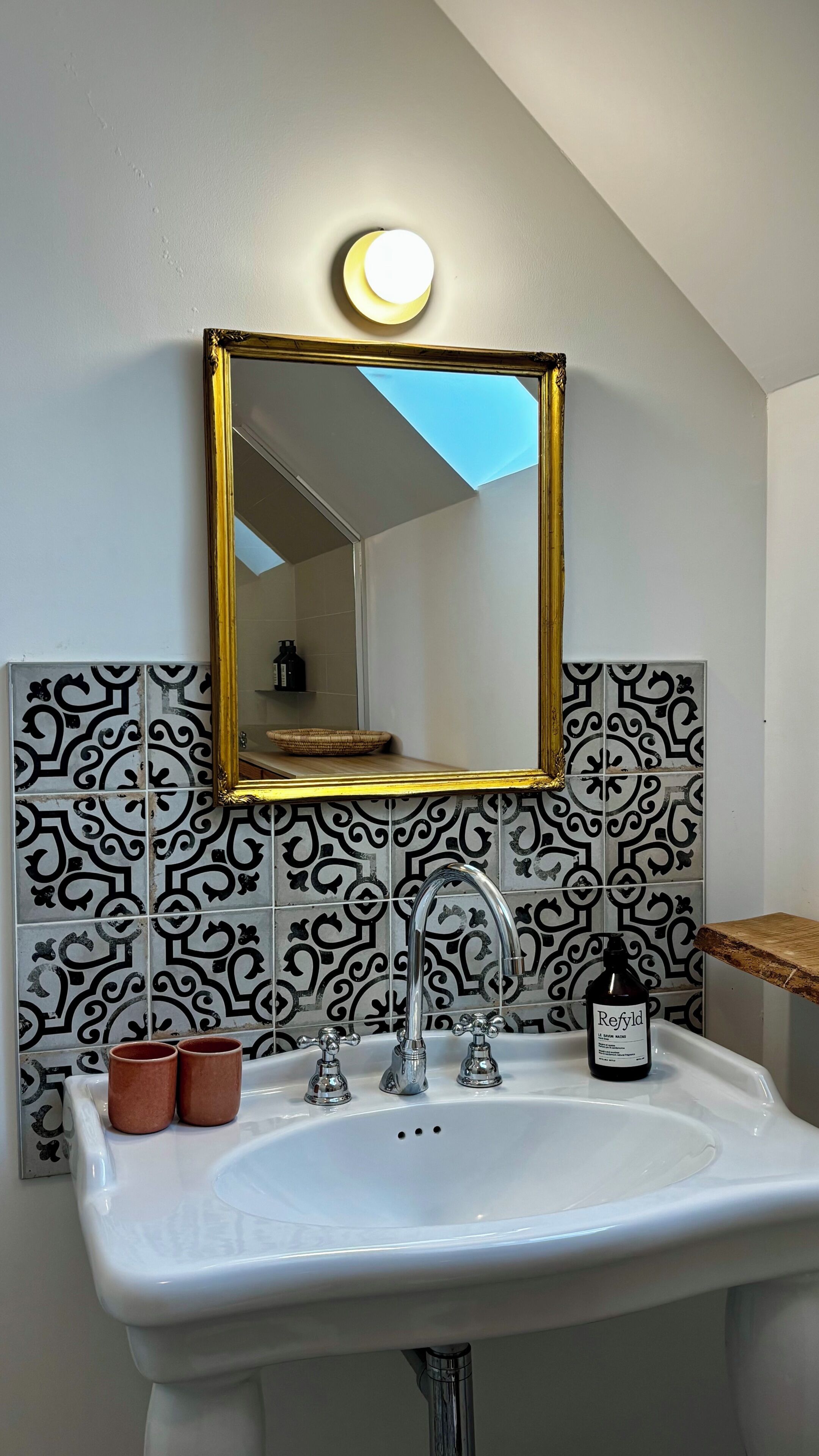 Ein Badezimmer mit einem weißen Waschbecken, einem spiegel mit goldener Rahmung und gefliesten Wänden.