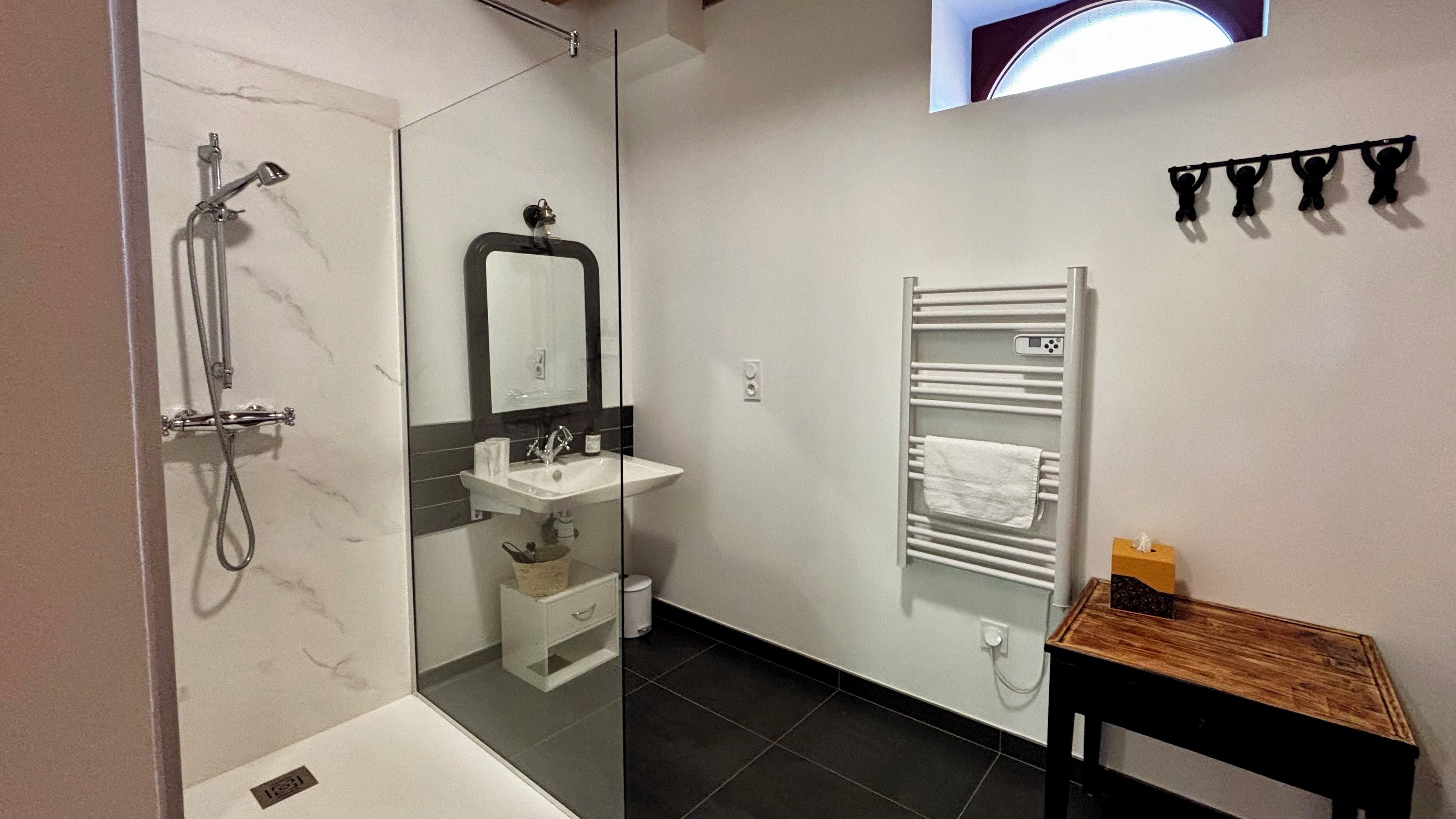 A modern bathroom with a glass-enclosed shower, a white sink, a mirror, a towel rack, and a wooden bench.