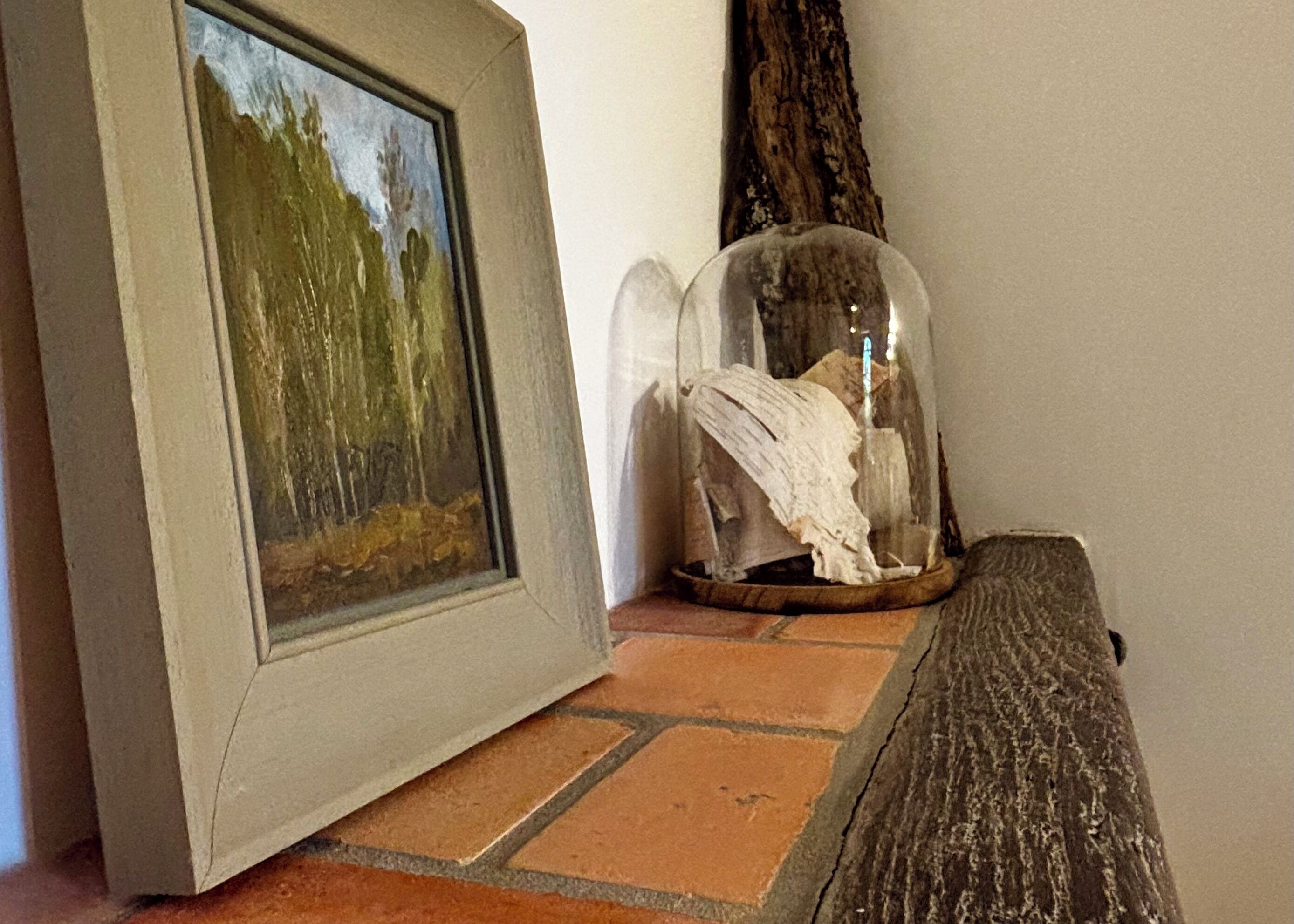 Un tableau encadré représentant une scène de forêt, une cloche en verre contenant un objet blanc, et une surface en bois.